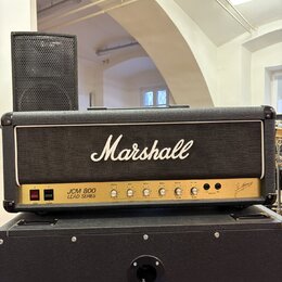 Kytarový kombozesilovač značky Marshall, model JCM 800 LEAD SERIES. Má černý kryt z textilie s bílým logem Marshall a ovládací panel s několika zlatý…