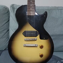 Elektrická kytara s tělem v žlutém gradientu (sunburst) a černým pickguardem. Nástroj má jeden humbucker v pozici mostu, dva zlaté ovládací knofíky a…