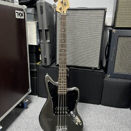 Tmavě šedá elektrická basová kytara s černým pickguardem a světle hnědým hmatníkem. Na hlavě nástroje je nápis JAGUAR BASS a logo Fender. Kytara stoj…