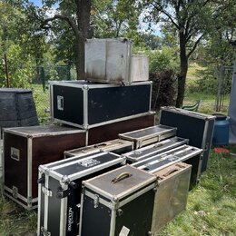Skupina transportních case různých velikostí a barev (černé, hnědé, stříbrné) umístěná na trávníku. Na jedné z černých case je viditelný nápis ACCUSR…