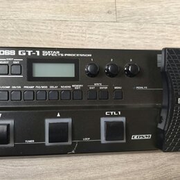 Černý kytarový procesor s nápisem BOSS GT-1 GUITAR EFFECTS PROCESSOR. Zařízení obsahuje displej, tři ovládací knoflíky, řadu tlačítek a dvastředý ped…