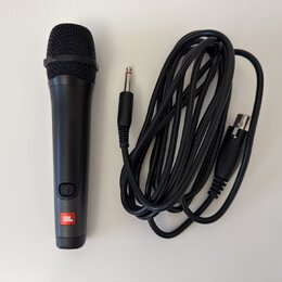 Černý dynamický mikrofon s mřížkou a připojeným černým kabelem. Na spodní části mikrofonu je umístěno červeno-bílé logo JBL a nad ním se nachází tlač…