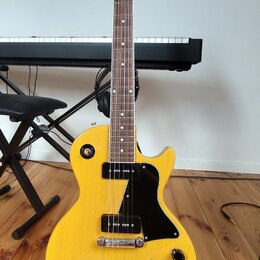 Žlutá elektrická kytara typu Les Paul s černým pickguardem a černými knoflíky. Na hlavě hmatníku je bílý nápis "Tokai". Nástroj je umístěn na černém…