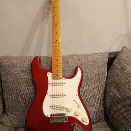 Elektrická kytara s červeným trupem, bílým pickguardem a hnedým javorovým hmatníkem. Na hlavě je viditelné logo Fender. Kytara má tři snímače typu si…