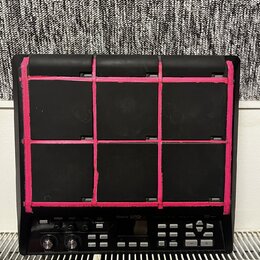 Černý elektronický sampling pad s devíti dopadovými polmi. Mezi polmi jsou nalepené výrazné růžové pásky. Na spodním panelu je bílý nápis Roland SPD-…