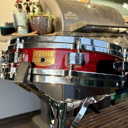 Červený snare drum s chromovanými obručmi a ladicími šrouby. Na korpusu je zlatý nápis TAMA a štítek s textem SNARE DRUM, LEGAL No. 200, MADE IN JAPA…