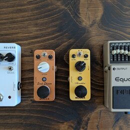 Čtyři kytarové efekty v řadě na dřevěném povrchu. Zleva: bílý pedál s nápisy REVERB, JHS 3 SERIES a ovládacími prvky VERB, EQ, DECAY a PRE DELAY. Dru…