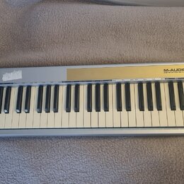 Stříbrný MIDI kontrolér s klaviaturní klávesou. Na horním panelu je zlatý pruh a nápis M-AUDIO KEYSTATION 61. V levé části jsou umístěny ovládací kno…