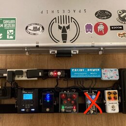 Stříbrný hliníkový kufr s různými nálepkami, včetně nápisů SPACESHIP, TONERIDER a KingTone. Pod kufrem je černý pedalboard s napájecím trafem Caline…