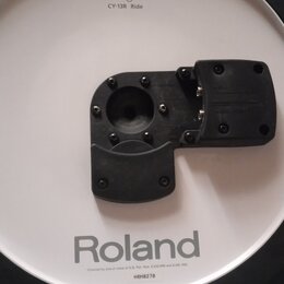 Bílý elektronický činel s černým gumovým okrajem a černým plastovým úchytem v centru. Na povrchu jsou nápisy V-Drums, CY-13R Ride a Roland. Dole je u…