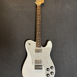 Bílá elektrická kytara s hnědým hmatníkem a perleťovým pickguardem. Na hlavě je viditelné logo Fender a nápis Telecaster Deluxe. Nástroj vykazuje zná…