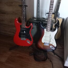 Na fotografii jsou čtyři kytary v držácích a jeden černý kombo zesilovač. V popředí jsou dvě elektrické kytary typu Stratocaster, jedna v červené bar…
