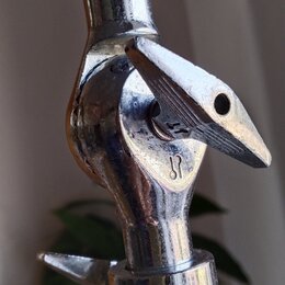 Detail chromovaného spojovacího kloubu stativu s černým knoflíkem nahoře. Na kovové části je vyryté logo připomínající stylizované písmeno S. Povrch…