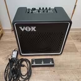Černý kytarový kombozesilovač s bílým rámečkem kolem mřížky a logem VOX. Na horním ovládacím panelu jsou umístěny ovratidla a přepínače. Před zesilov…