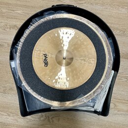 Dvě mosazné činely v černém plastovém kufru, přikryté průhlednou fólií. Na horním činele je viditelné logo Paiste. Mezi činely je umístěna černá tlum…