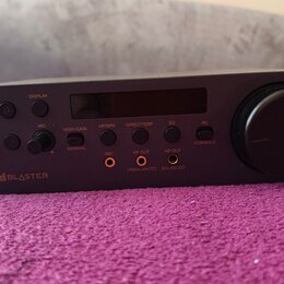 Tmavě šedý audio interface s digitálním displejem a velkým ovládacím knoflíkem vpravo. Na předním panelu je nápis Sound BLASTER a několik tlačítek s…