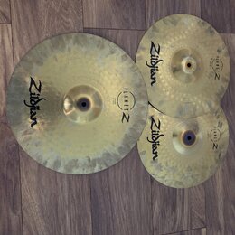 Tři mosazné činely v různých velikostech položené na hnědém laminátovém povrchu. Každý činel nese černý nápis Zildjian a logo PLANET Z. Povrchy činel…