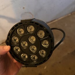 Černý kulatý LED reflektor s 12 světelnými zdroji rozmístěnými v kruhu. Zařízení má plastové tělo a integrovanou úchytnou rukojeť. Na fotce nejsou vi…