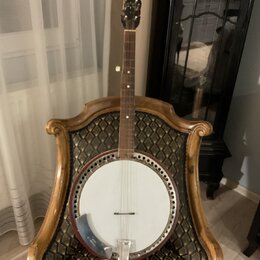 Banjo s bílou membránou, hnědým dřevěným rímem a krčkem. Na hlavě nástroje je viditelné logo, které není čitelné. Nástroj je opřen o zdobenou židli s…
