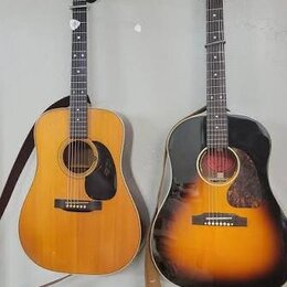 Dvě akustické kytary zavěšené na stěně. Levá kytara má světlý dřevěný povrch, černý pickguard a hnědý popruh. Pravá kytara má povrch v provedení sunb…