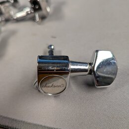 Detailní pohled na jeden chromovaný ladicí kolík kytary. Na kulaté části s nápisem je viditelné logo Jackson®. Povrch vykazuje drobné škrábance a zná…