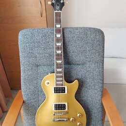 Zlatá elektrická kytara typu Les Paul s dvěma humbuckery a čtyřmi ovládacími knoflíky. Na hlavě je bílý nápis Epiphone a zlatý nápis Les Paul. Kytara…