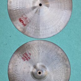 Dvě kovové činely v stříbrné barvě s viditelným broušením a otisky kladiva. Horní činel nese nápisy HEAVY HI-HAT BOTTOM 14" a logo Paiste 2002. Dolní…
