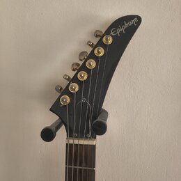 Fotografie zachycuje hlavu elektrické kytary v černé barvě. Na špičce hlavy je bílý nápis Epiphone. Nástroj má šest zlatých ladiček a hnedík z tmavéh…
