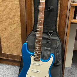 Modrá elektrická kytara s bílým pickguardem a hnědým hmatníkem, opřená o černý pouzdro. Na hlavě kytary je viditelné logo Telecaster. Nástroj má tři…
