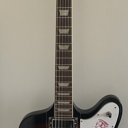 Elektrická kytara s tmavě hnědým gradientním lakem a bílým plechtrátem. Na hlavě je zlatý nápis Epiphone a na plechtrátu se nachází červené logo. Nás…