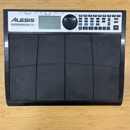 Černý elektronický bicí pad s osmi gumovými polštáři v rozložení 2x4. V horní části je bílý ovládací panel s nápisem ALESIS a označením PERFORMANCE P…