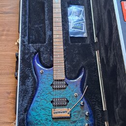 Elektrická kytara s modře gradientním tělem z plaménkového javoru, umístěná v černém tvrdém kufru. Nástroj má tři humbuckery, hned dva ovládací knofí…