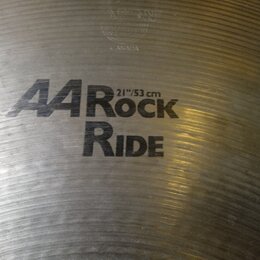 Detailní pohled na kovový činel s viditelným broušením povrchu. Na čineli je černý nápis „A4 Rock RIDE“ a pod ním označení velikosti „21"/53 cm“. Ve…