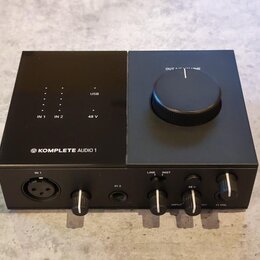 Černý zvukový interface s nápisem KOMPLETE AUDIO 1 a logem výrobce. Na horní straně jsou kontrolky s popisky IN 1, IN 2, USB a 48 V, vpravo je velký…
