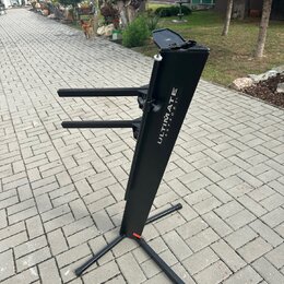 Černý kovový stojan na klávesové nástroje s dvěma vodorovnými rameny. Na hlavní nosné tyči je bílý nápis ULTIMATE SUPPORT™. Stojan stojí na dlážkovém…