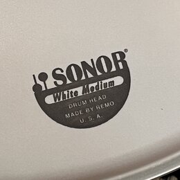Detail bílého bubnového koženu s černým potiskem. Je na něm vidět logo SONOR, nápis White Medium a text DRUM HEAD MADE BY REMO U. S. A.. Povrch je hl…