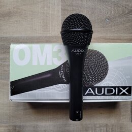 Černý dynamický mikrofon s mřížkou, na těle kterého je bílý nápis AUDIX OM3. Mikrofon leží na originální krabici, na které jsou vidět nápisy OM3 a AU…