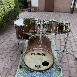 Bubnová sada v tmavě hnědé dřevěné povrchové úpravě. Na basovém bubnu je bílá kožená blana s nápisy SONOR a SQ2 DRUM SYSTEM. Na ostatních bubnech jso…