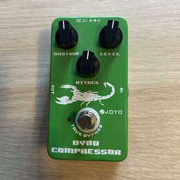Zelený kytarový efekt s nápisy DYNA COMPRESSOR a JOYO. Na horní straně jsou tři černé otočné ovířky s popisky SUSTAIN, ATTACK a LEVEL. Uprostřed je b…
