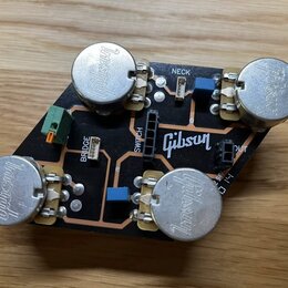 Černá tiskovaná spoje s четыřmi stříbrnými kovovými potenciometry, na kterých je vyražen nápis Gibson. Na desce jsou bílé nápisy BRIDGE, NECK, SWITCH…