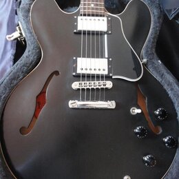 Černá polodupatá elektrická kytara s černým pickguardem a dvěma humbuckery. Nástroj je umístěn v otevřeném kufru s šedým polstrováním. Na těle jsou č…
