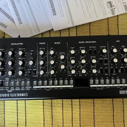 Černý analogový syntetizátor s nápisem SE-02 STUDIO ELECTRONICS a logem Roland. Přední panel obsahuje numeroustočítka, přepínače a sekci s klávesovým…