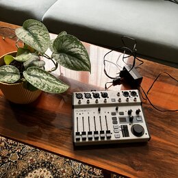 Na dřevěném stole je umístěn kompaktní mixážní pult stříbrné barvy s černými ovládacími prvky. Na zařízení je viditelné logo Behringer a nápis FLOW 8…