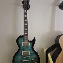 Elektrická kytara s tmavě zeleným gradientem a viditelnou strukturou dřeva. Na hlavě je bílý nápis Cort. Nástroj má dva humbuckery, čtyři otočné knof…