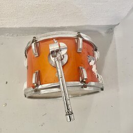 Oranžový buben s kovovými obručemi a lugy. Na korpusu je vidět logo s nápisem DUGGERY, které je v pravé části částečně odloupané a poškozené. K bubnu…