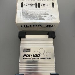 Fotografie zobrazuje stříbrnou kovovou pasivní DI box s černými plastovými zátlačkami na koncích. Na horní straně je nápis DAP AUDIO a model PDI-100…