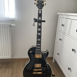 Černá elektrická kytara s gold hardwarem a bílým bindingem, umístěná na stojanu. Na hlavě hmatníku je bílý nápis Epiphone. Kytara má dva humbuckery,…