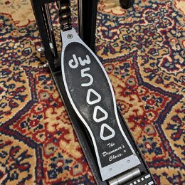 Černý bicí pedál s kovovými prvky. Na šlapadle je bílým nápisem značka dw a model 5000, dále text The Drummer's Choice.® a logo dw na horní části kon…