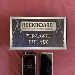 Na fotografii je krabička a dvě součásti příslušenství. Na krabici je nápis ROCKBOARD® by WARWICK a ručně napsaný text PEDALSAFE PICK BOX. Vedle krab…
