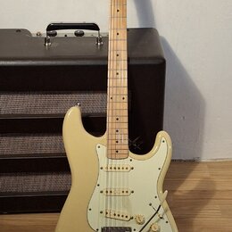 Krémová elektrická kytara typu Stratocaster s bílým pickguardem a javorovým hmatníkem. Na hlavě je viditelné logo Squier. Kytara je umístěna před čer…